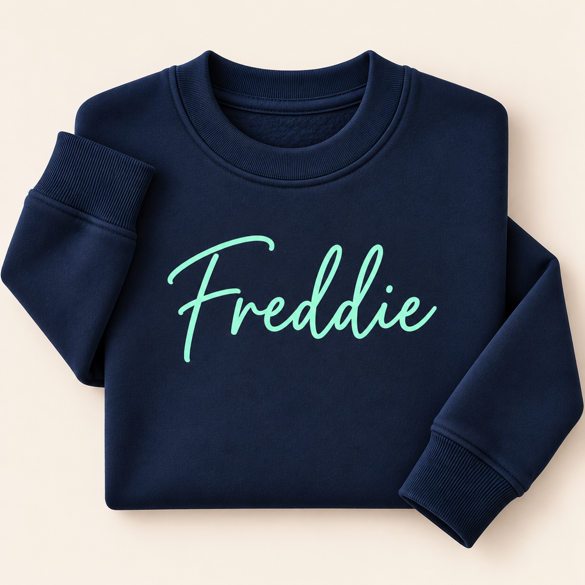 Navy personalised sweatshirt with a mint green script name across the chest, folded on a beige background | Grace & Grayson