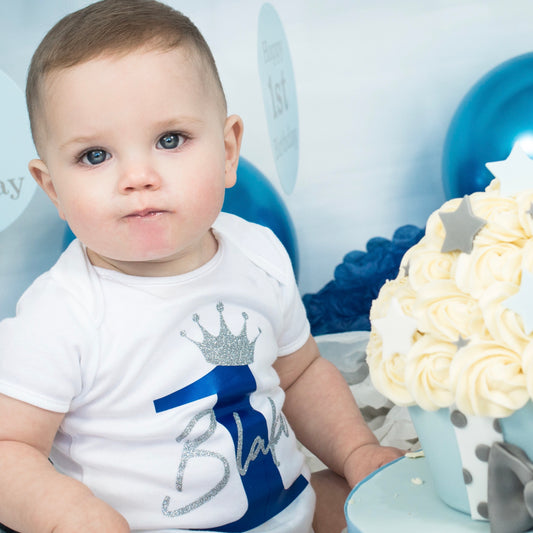 Baby boy wearing personalised Birthday Number 1 baby vest with blue pearl and silver glitter crown design at cake smash - Grace & Grayson