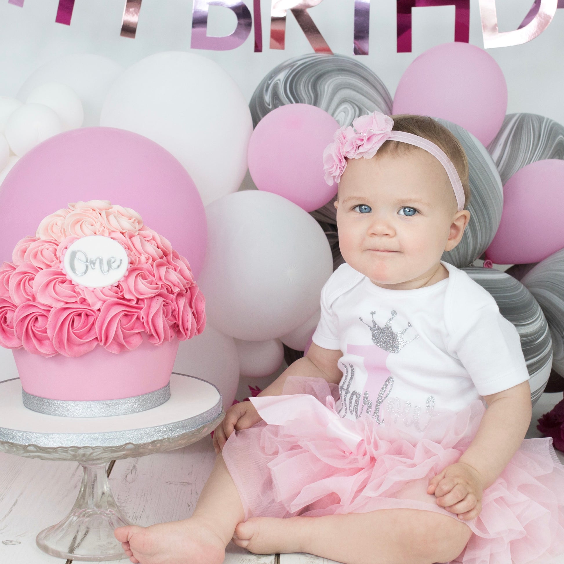 Baby girl in personalised birthday tutu set with pink tutu and silver crown vest sitting next to birthday cake at cake smash photoshoot - Grace & Grayson