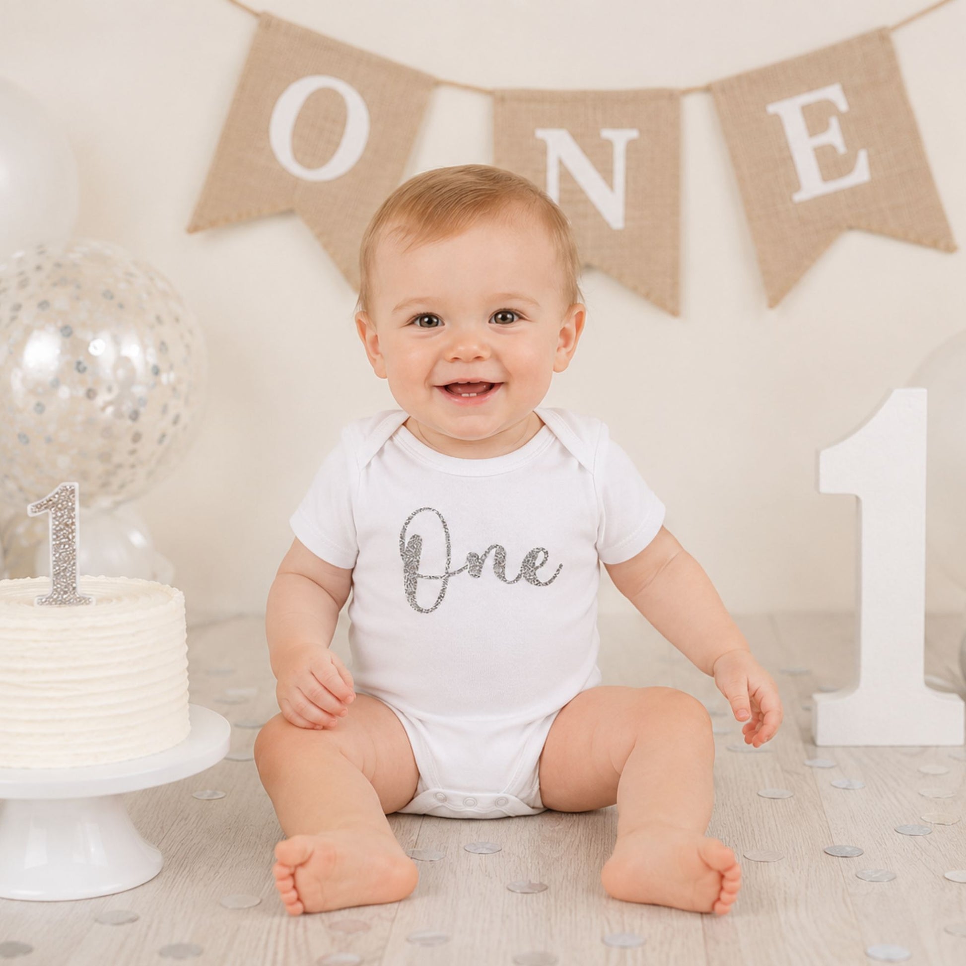 Baby wearing personalised 1st birthday baby vest with silver glitter One design, sitting next to birthday cake at first birthday party - Grace & Grayson