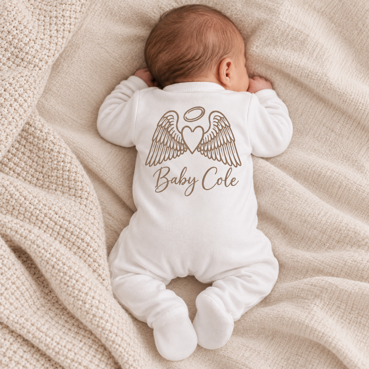 Baby wearing white footed baby grow with a large personalised angel wings and halo print in tawny brown, laid flat against a pale background, Grace & Grayson