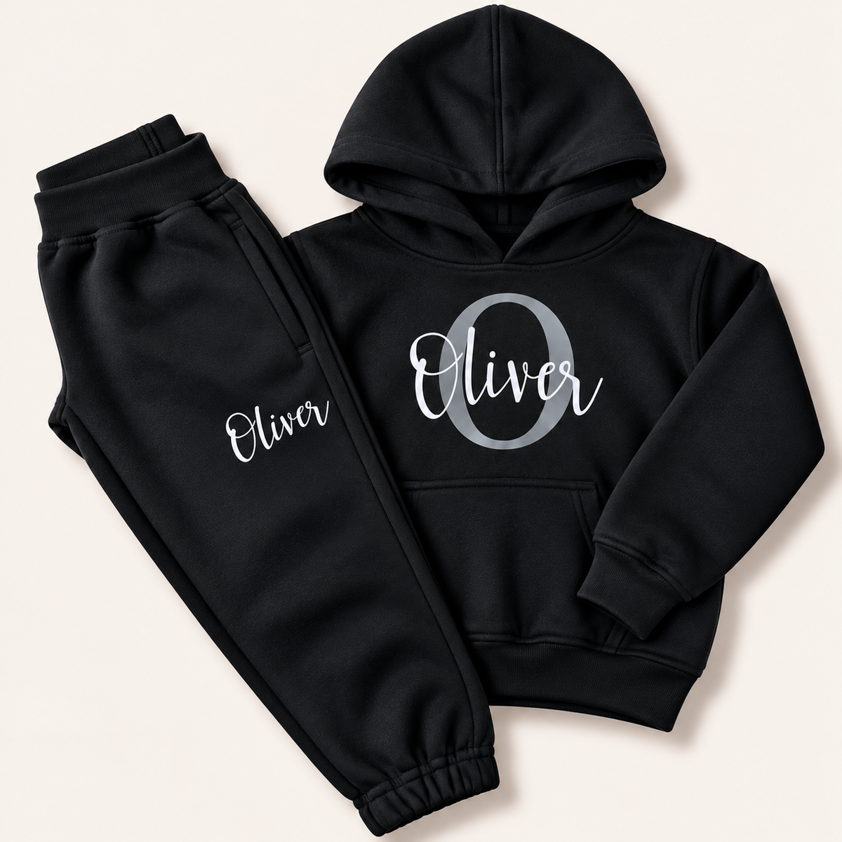 Black hoodie and joggers with a grey initial and white script name personalisation, laid flat on a cream background. Grace & Grayson