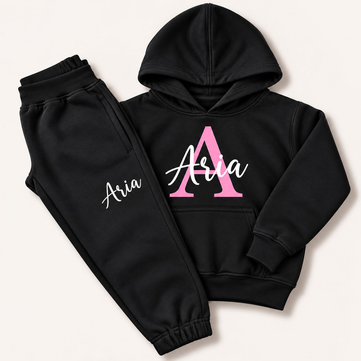 Black hoodie and joggers with a pink initial and white script name personalisation, laid flat on a cream background. Grace & Grayson