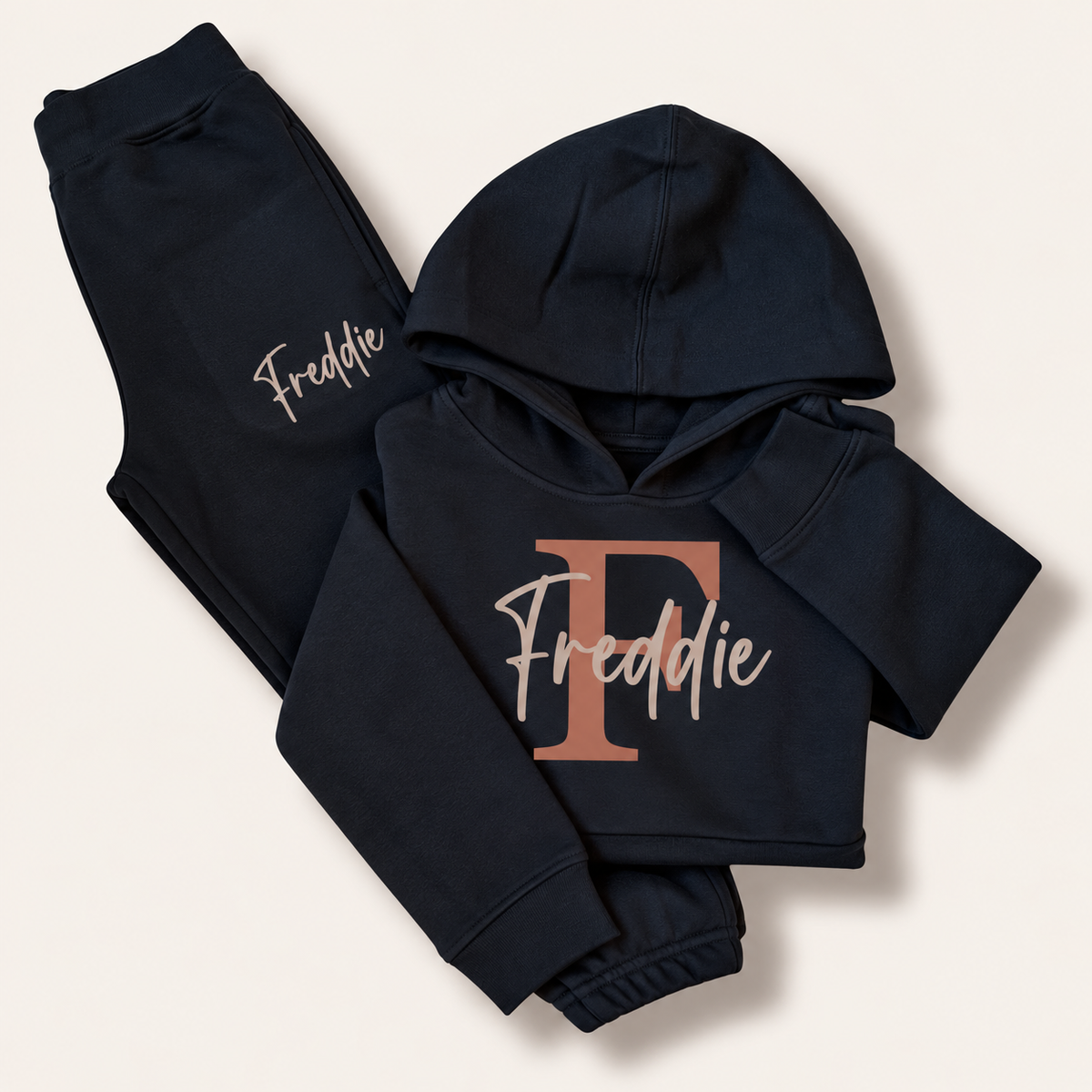 Black folded hoodie tracksuit with a taupe initial and script name personalisation, laid flat on a cream background. Grace & Grayson