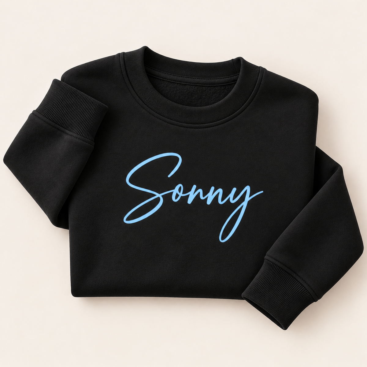 Black personalised sweatshirt with a light blue script name across the chest, folded on a beige background | Grace & Grayson