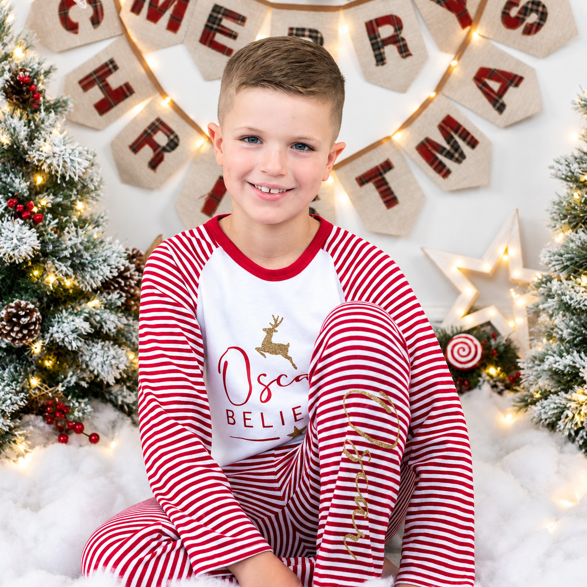 Child model wearing red and white striped personalised Believes Christmas pyjamas in a festive Christmas setting, Grace & Grayson