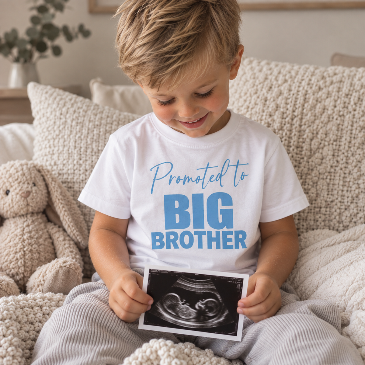 Child wearing a white “Promoted to Big Brother” t-shirt and holding an ultrasound photo, Grace & Grayson.