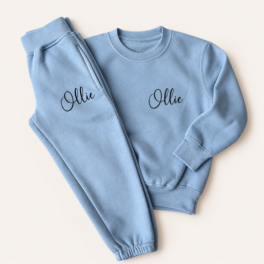 Dusty blue personalised kids tracksuit laid flat with a navy script name on the sweatshirt chest and jogger pocket, Grace & Grayson