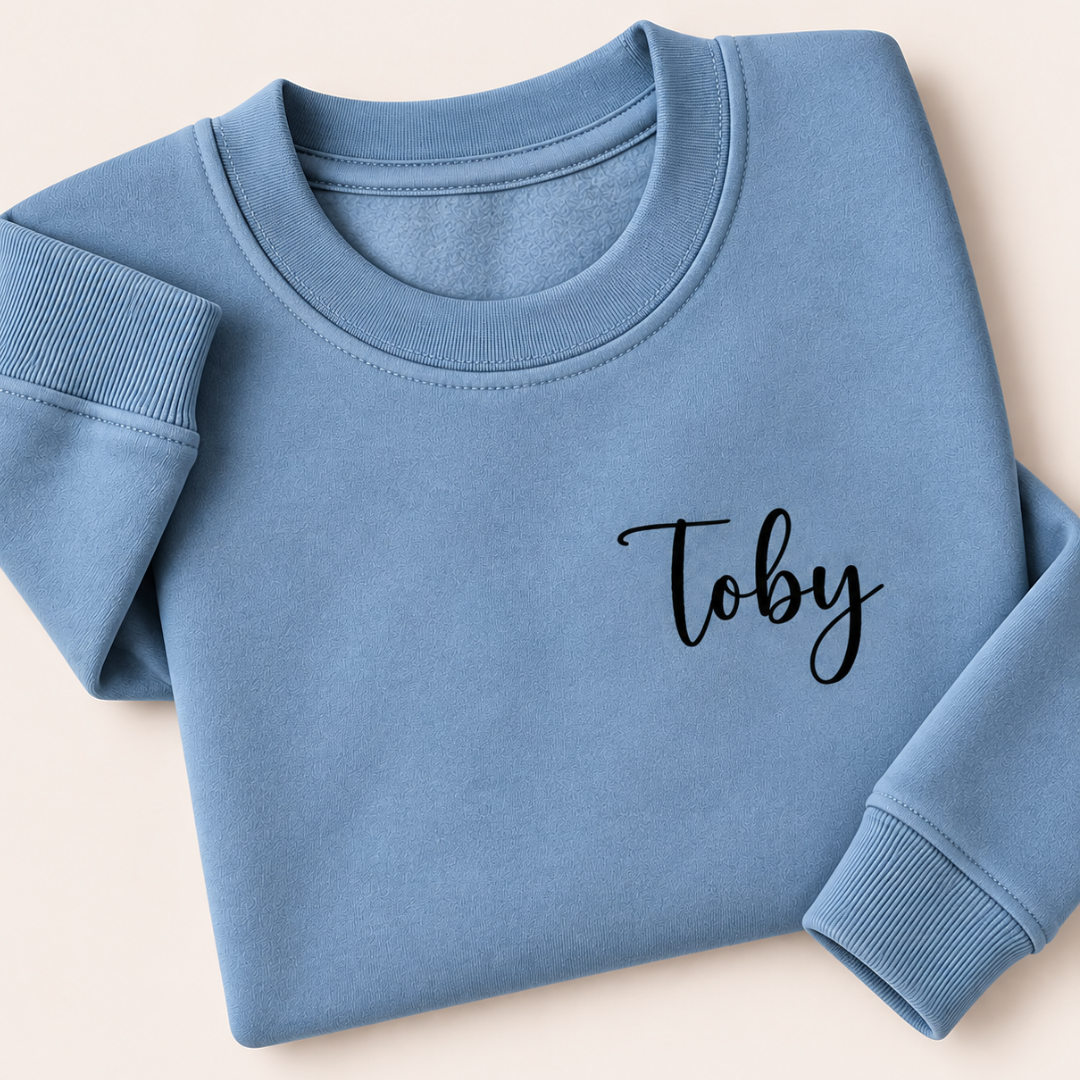 Dusty blue personalised sweatshirt with a small black script name on the chest, folded on a beige background | Grace & Grayson
