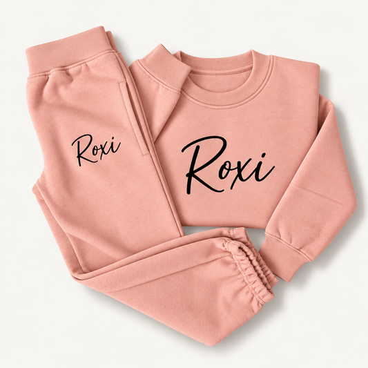 Dusty pink personalised tracksuit laid flat with a black script name across the sweatshirt chest and on the joggers, Grace & Grayson