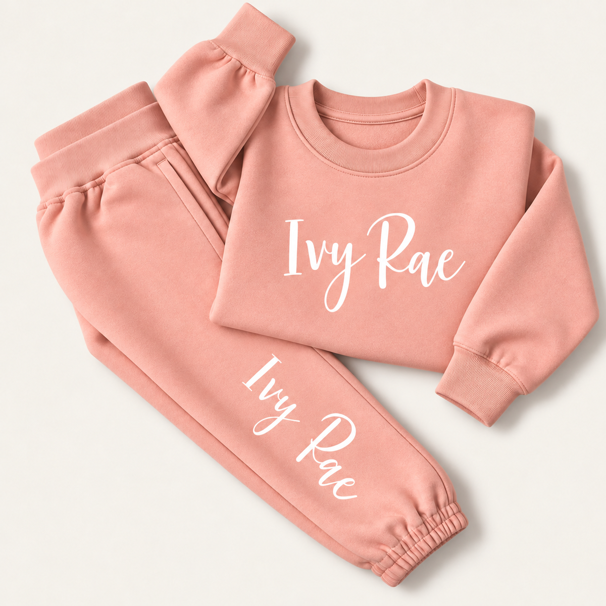 Dusty pink personalised tracksuit laid flat with a white script name across the sweatshirt chest and down the jogger leg, Grace & Grayson