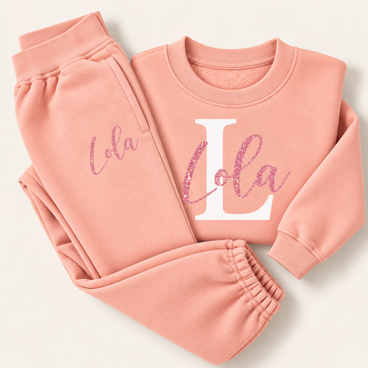 Dusty pink personalised sweatshirt and joggers laid flat with a pink glitter script name over a large white initial on the chest and a small name on the jogger pocket, Grace & Grayson
