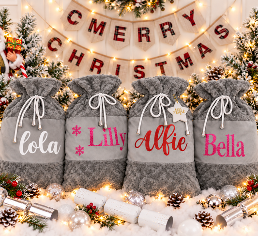 Four grey plush Christmas Santa sacks with white drawstrings arranged beneath a festive banner and lights, Grace & Grayson.