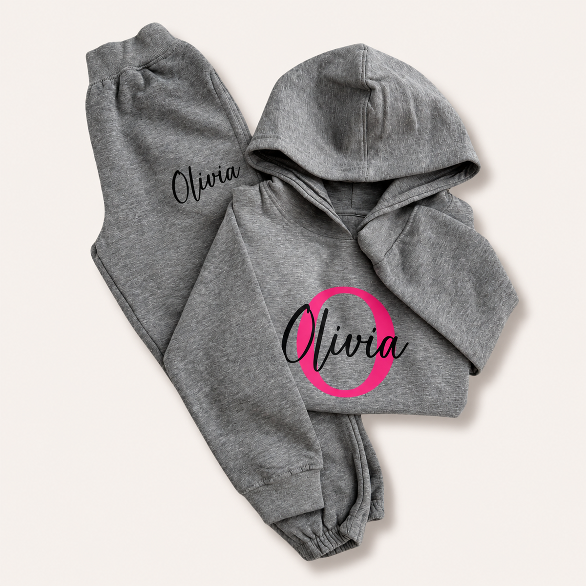 Grey hoodie tracksuit with a bright pink initial and black script name personalisation, laid flat on a cream background. Grace & Grayson