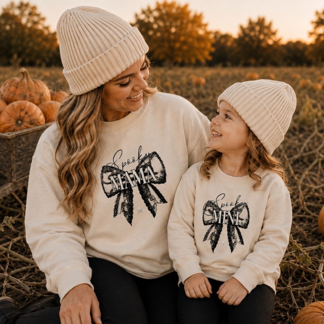 Mum and daughter wearing matching personalised Spooky Mama and Mini Halloween bow sweatshirts in oat, sitting in a pumpkin field by Grace & Grayson