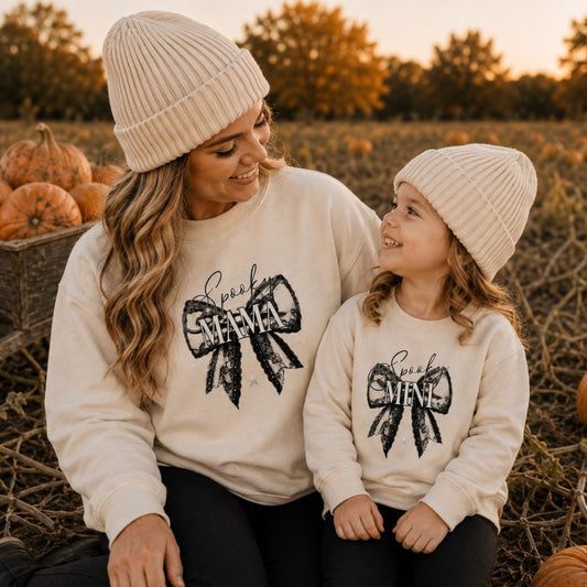 Mum and daughter wearing matching personalised Spooky Mama and Mini Halloween bow sweatshirts in oat, sitting in a pumpkin field by Grace & Grayson