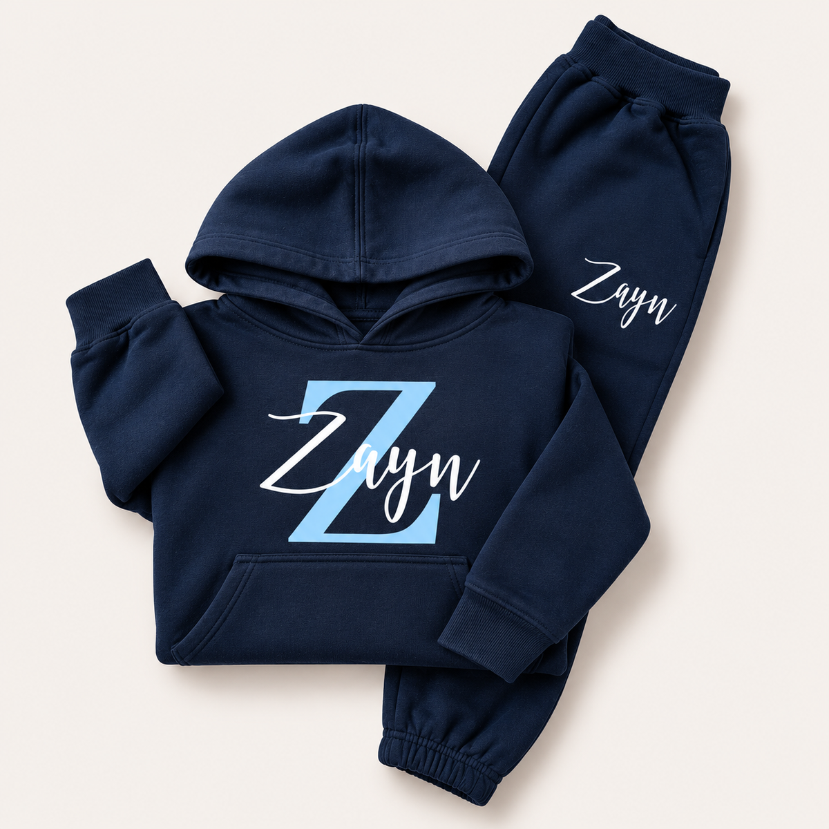 Navy hoodie tracksuit with a pale blue initial and white script name personalisation, laid flat on a cream background. Grace & Grayson