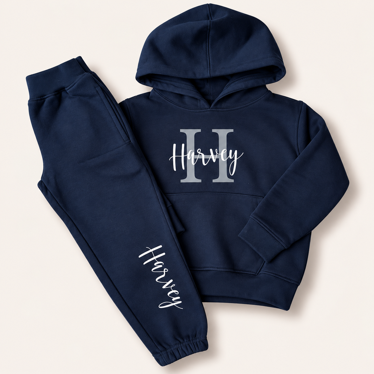 Navy hoodie and joggers with a grey initial and white script name personalisation, laid flat on a cream background. Grace & Grayson