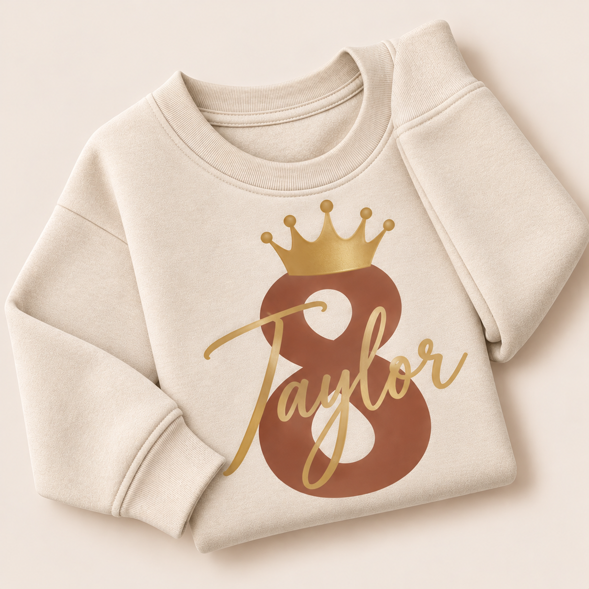 Oat personalised birthday sweatshirt with a brown number 8, gold crown and gold script name, folded on a beige background | Grace & Grayson