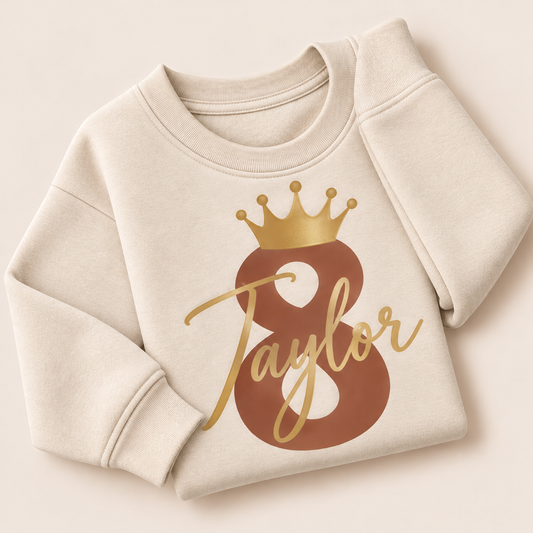 Oat personalised birthday sweatshirt with a brown number 8, gold crown and gold script name, folded on a beige background | Grace & Grayson