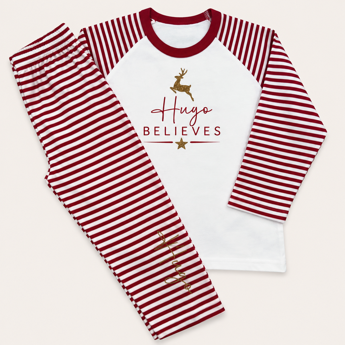 Red and white striped personalised Believes Christmas pyjamas with a gold Christmas tree design, silver BELIEVES lettering and personalised name, displayed flat, Grace & Grayson