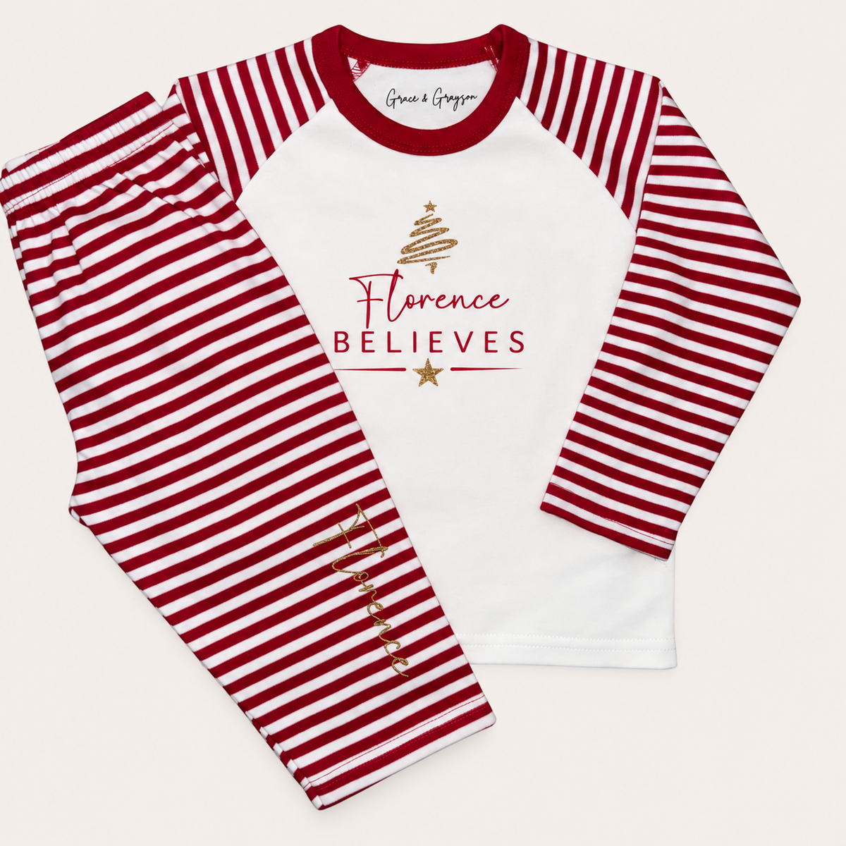 Red and white striped personalised Believes Christmas pyjamas with a gold christams tree design, red BELIEVES lettering and personalised name, displayed flat, Grace & Grayson