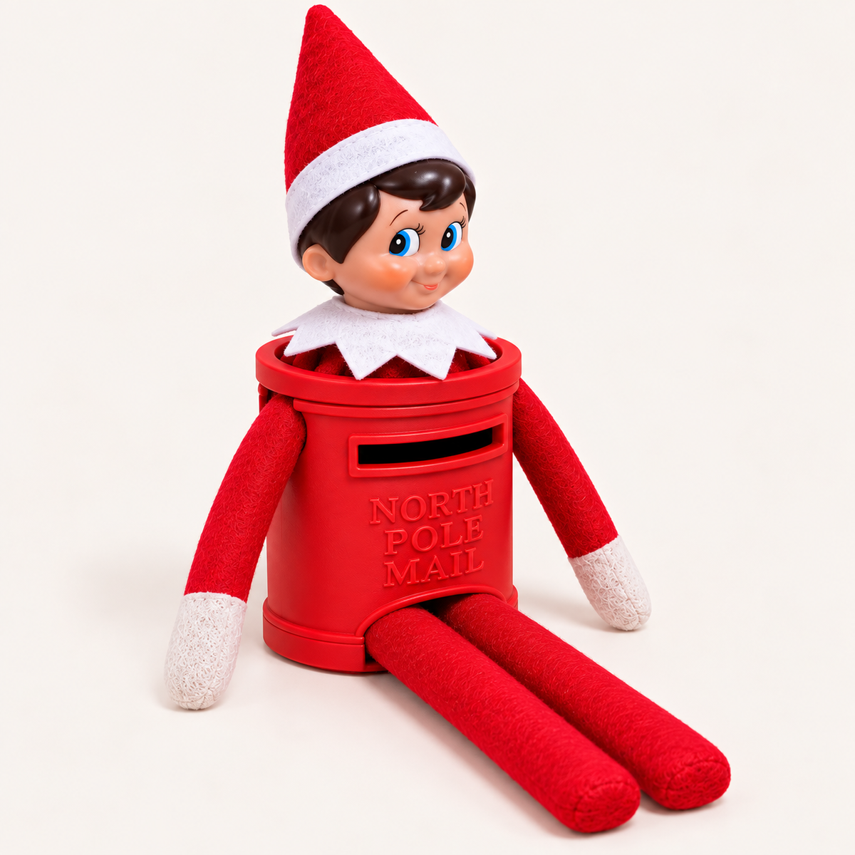 Christmas elf on the shelf seated at an angle in a red North Pole Mail post box costume against a light background.