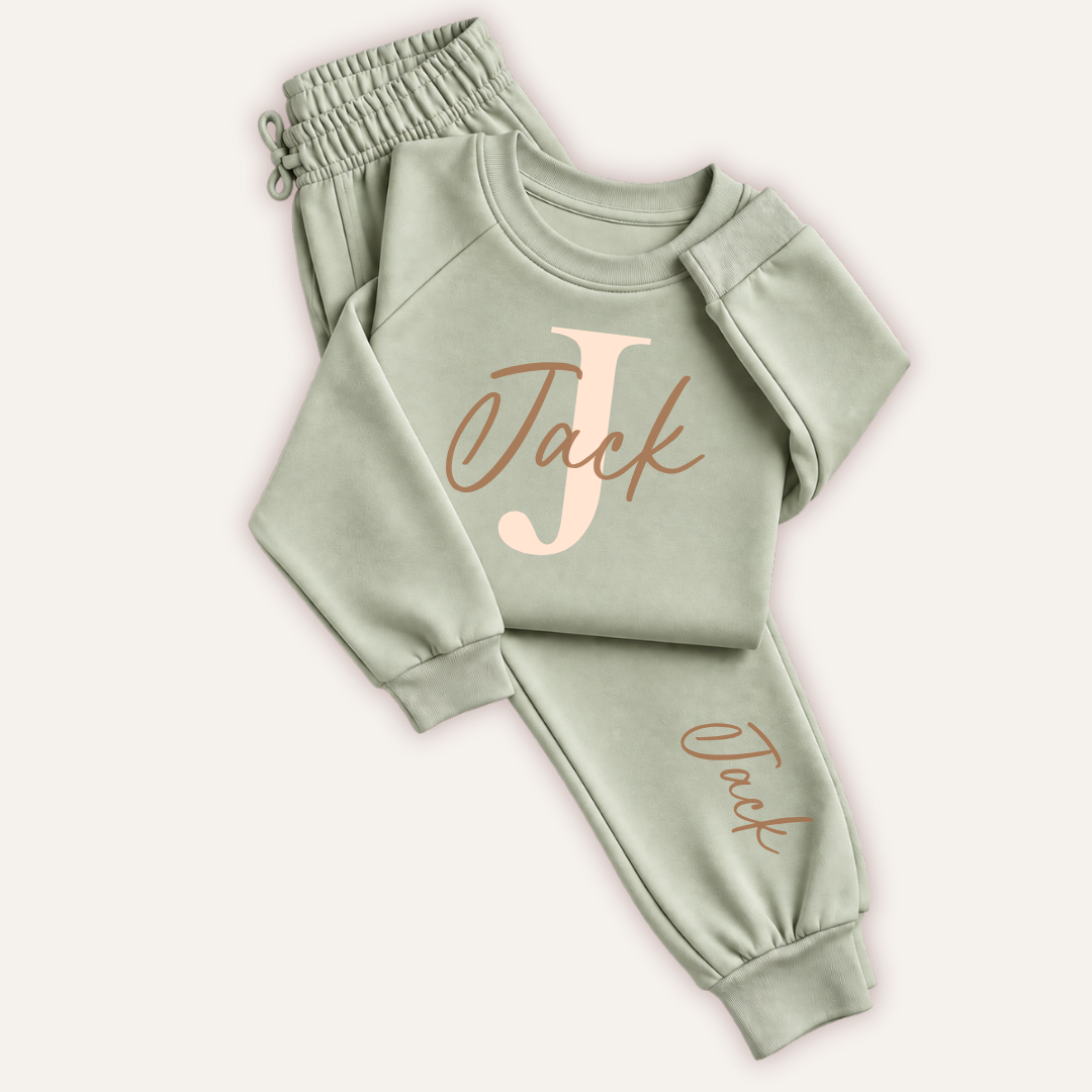 Sage green personalised children’s tracksuit laid flat on a light background, with a large initial and matching name on the sweatshirt and joggers.