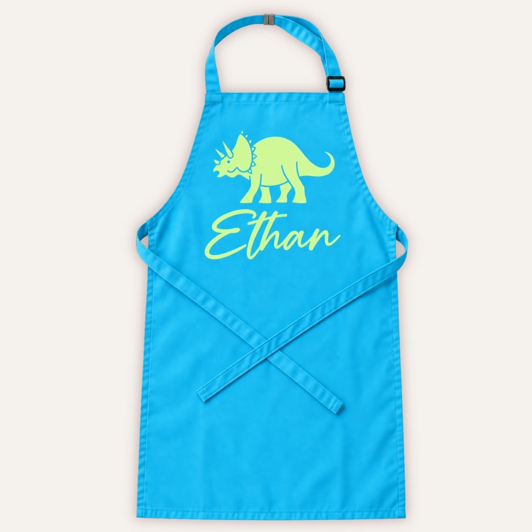Sky blue personalised children’s apron with pastel green dinosaur design on a white background, Grace & Grayson