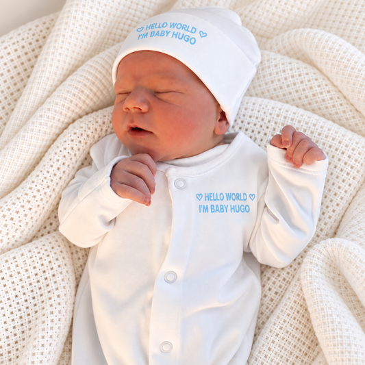 Sleeping newborn wearing a white personalised Hello World baby grow and matching hat with blue lettering, nestled in a cream knitted blanket, Grace & Grayson