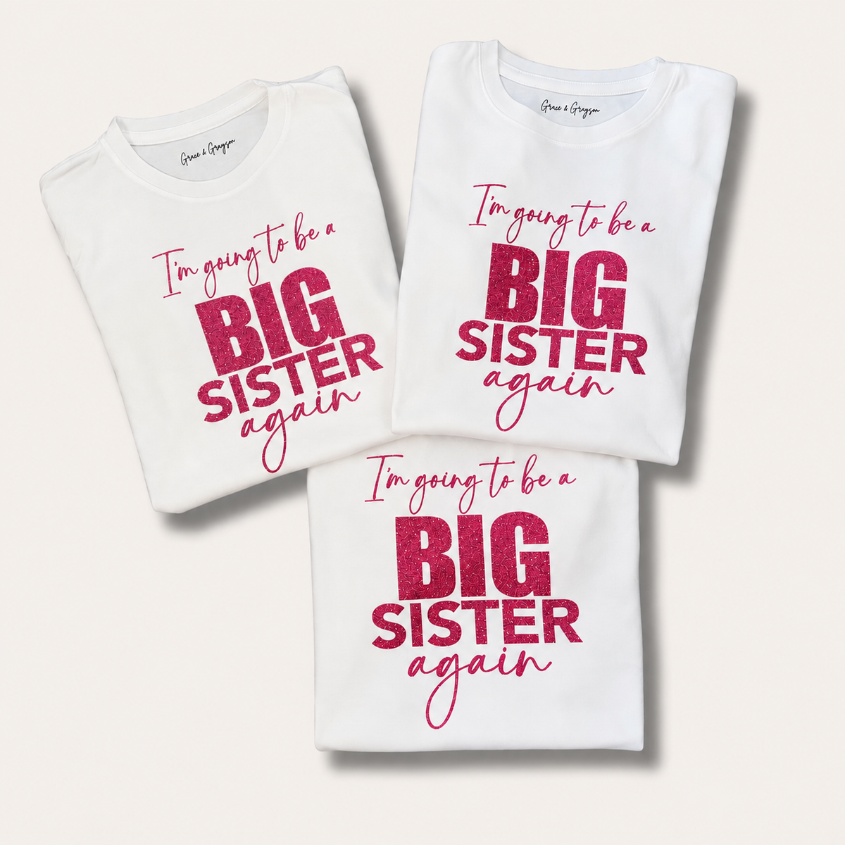 Three folded white personalised “I’m Going to Be a Big Sister Again” t-shirts, Grace & Grayson.