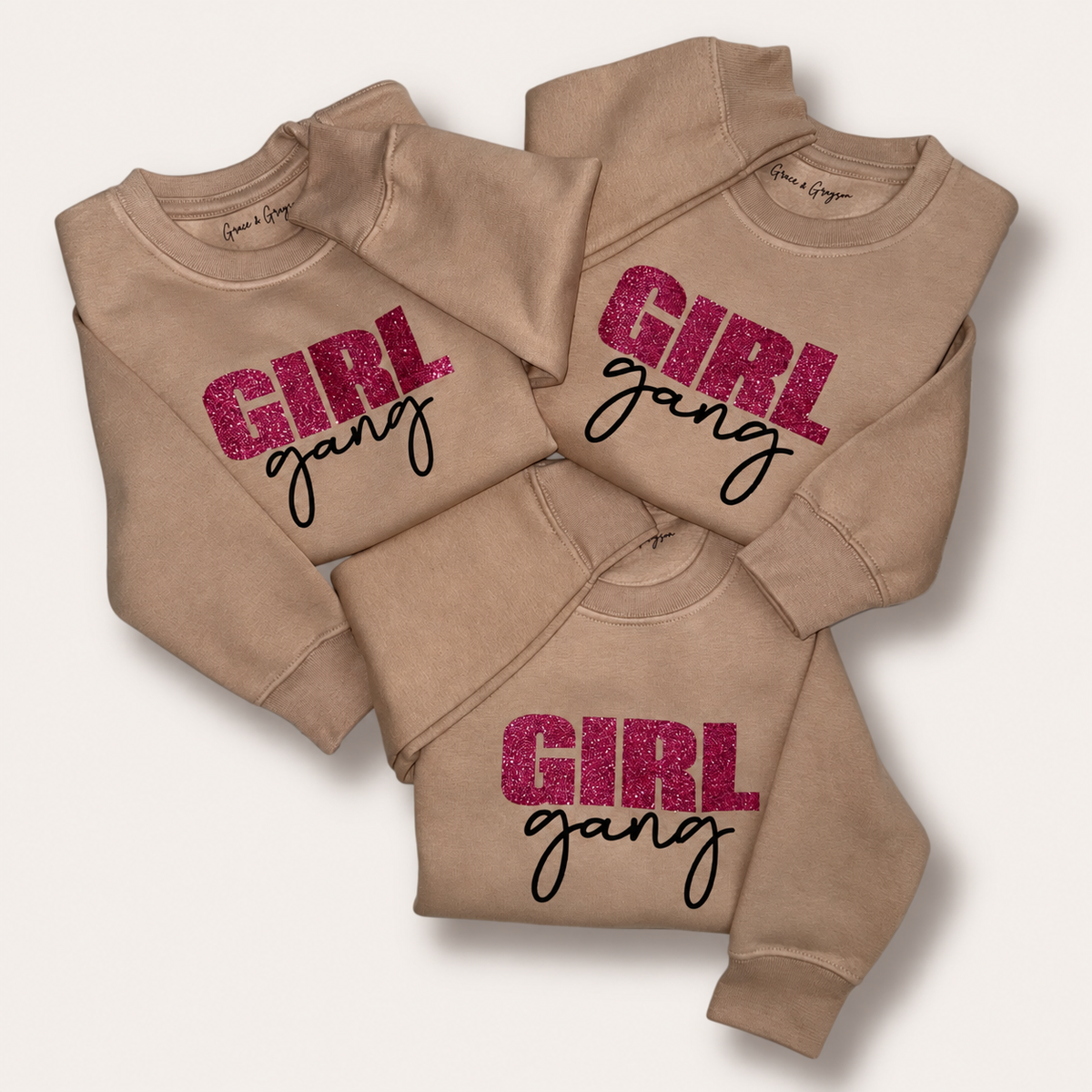 Three warm taupe Girl Gang sweatshirts arranged flat with pink glitter lettering and black script, Grace & Grayson