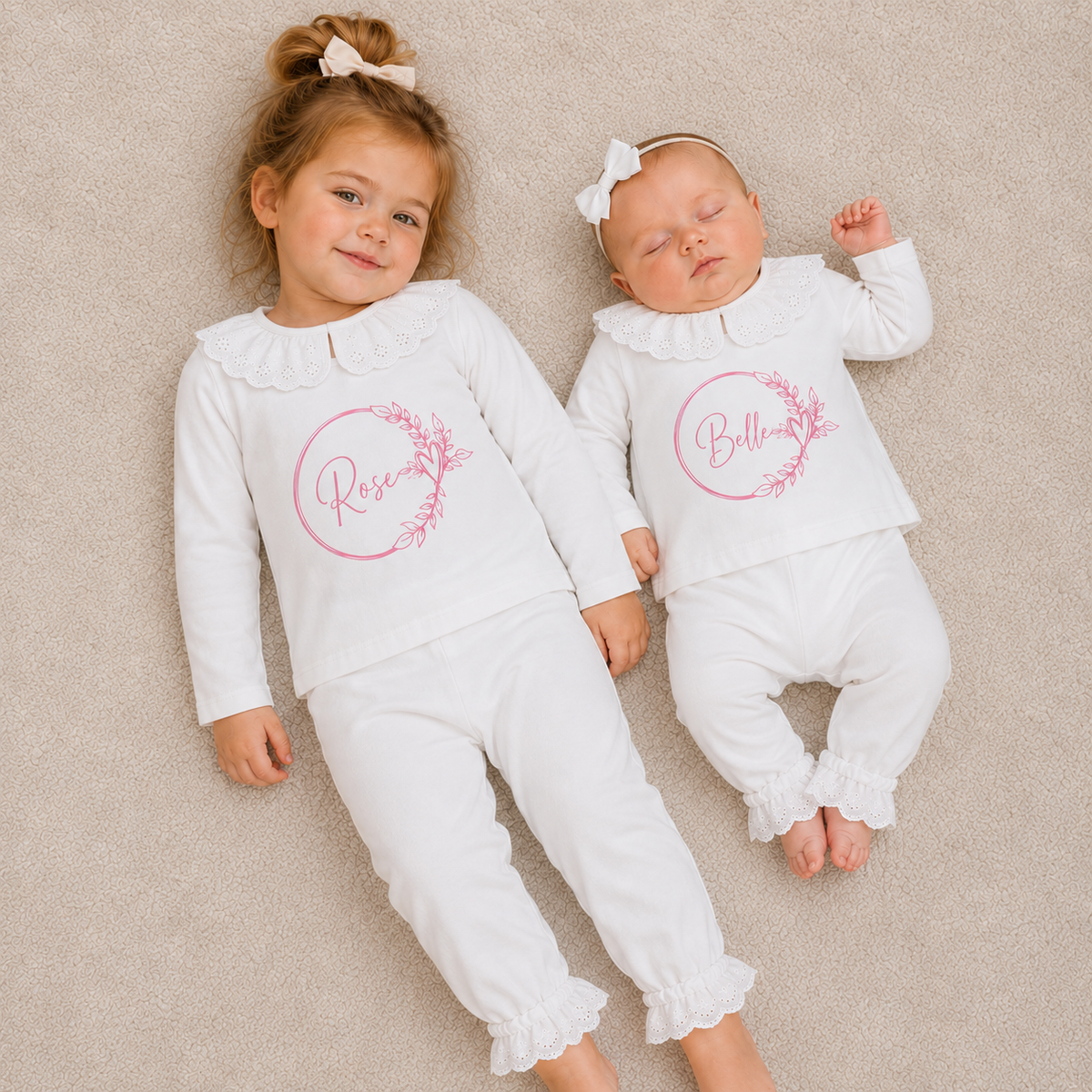 Two children wearing white personalised broderie frill pyjamas with pink wreath designs, lying on a beige carpet, Grace & Grayson
