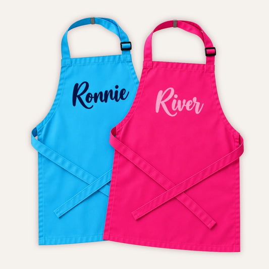 Two personalised children’s aprons, one blue and one pink, displayed on a white background, Grace & Grayson.
