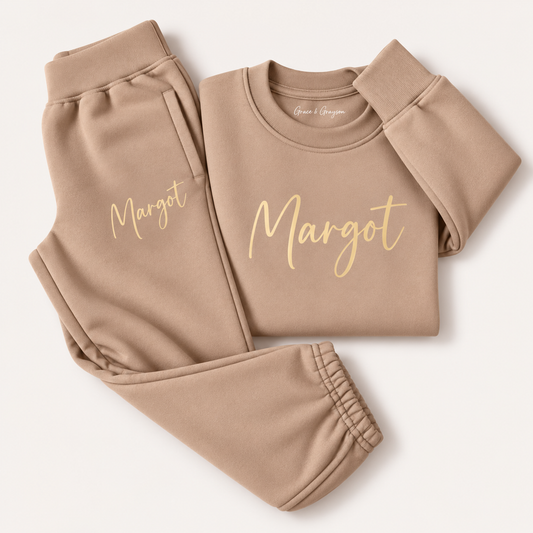 Warm taupe personalised kids tracksuit laid flat with a gold script name across the sweatshirt chest and on the jogger pocket, Grace & Grayson