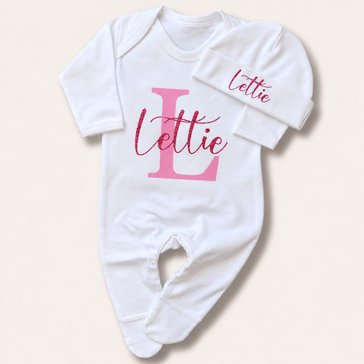 White personalised baby grow and matching hat with a pink glitter initial design, Grace & Grayson