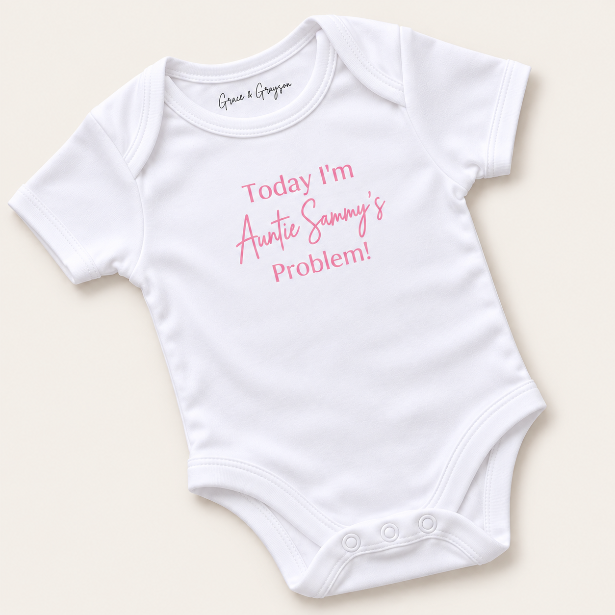 White personalised baby vest with pink lettering reading “Today I’m Auntie’s Problem!”, displayed at an angle on a light background, Grace & Grayson.