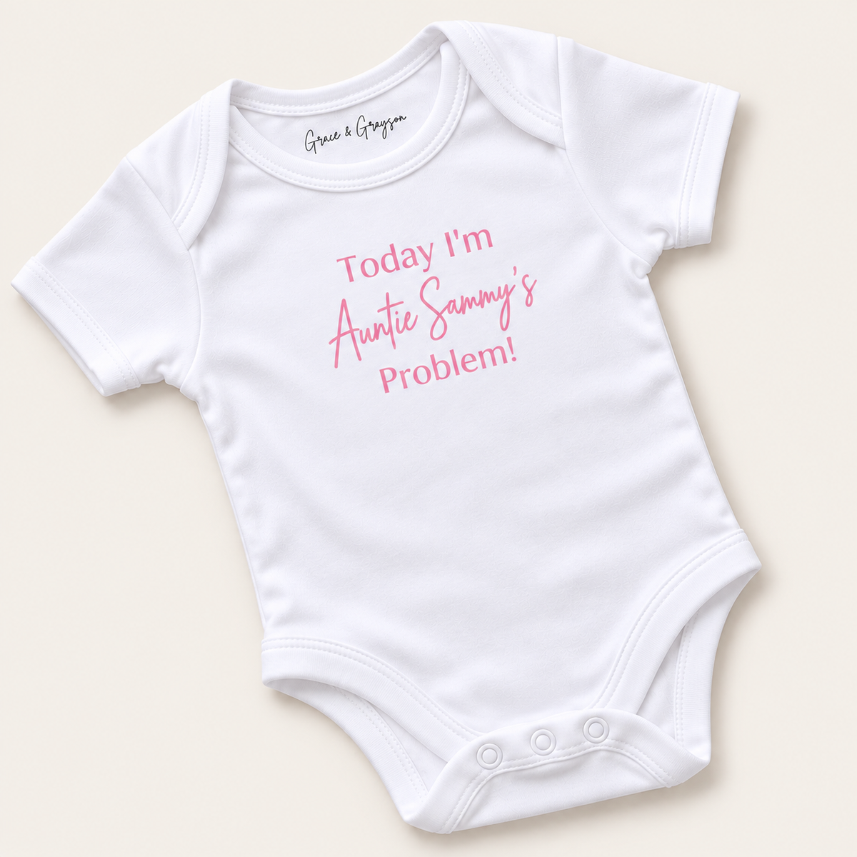 White personalised baby vest with pink lettering reading “Today I’m Auntie’s Problem!”, displayed at an angle on a light background, Grace & Grayson.