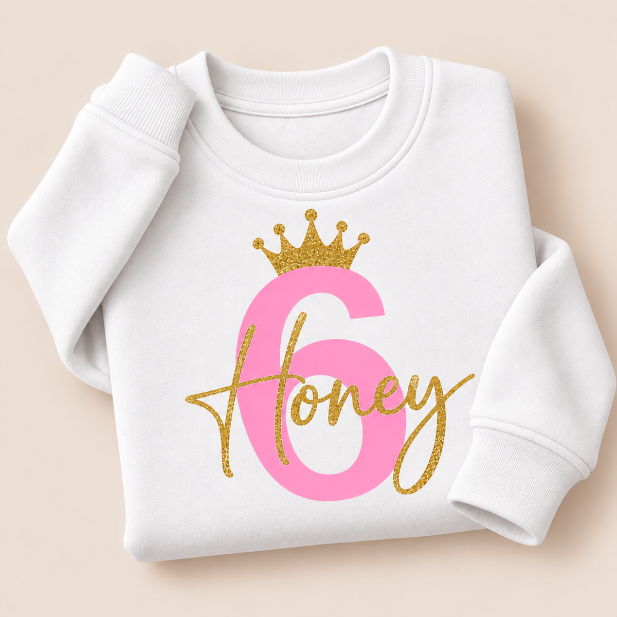 White personalised birthday sweatshirt with a pink number 6, gold glitter crown and script name, folded on a beige background | Grace & Grayson