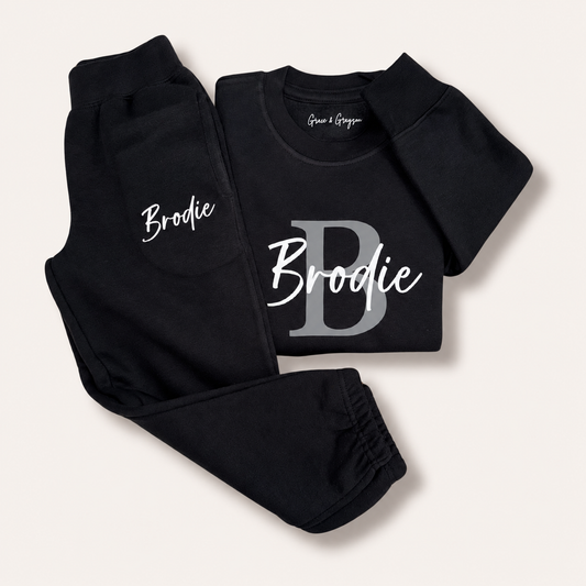 Black personalised sweatshirt and joggers with a grey initial and white script name, laid flat, Grace & Grayson