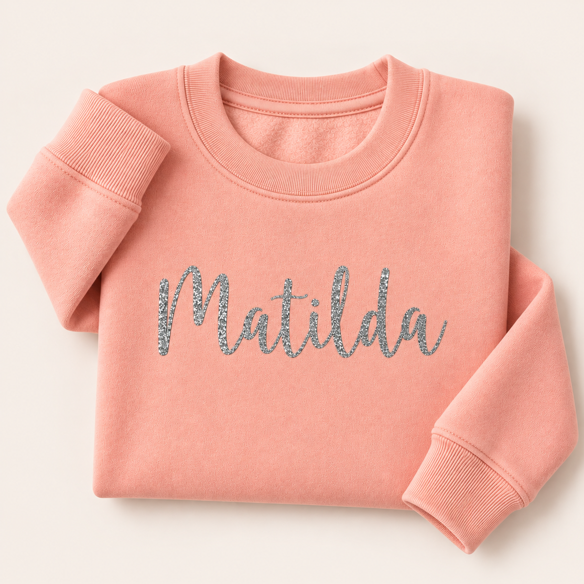 Peach personalised sweatshirt with a silver glitter script name across the chest, folded on a beige background | Grace & Grayson