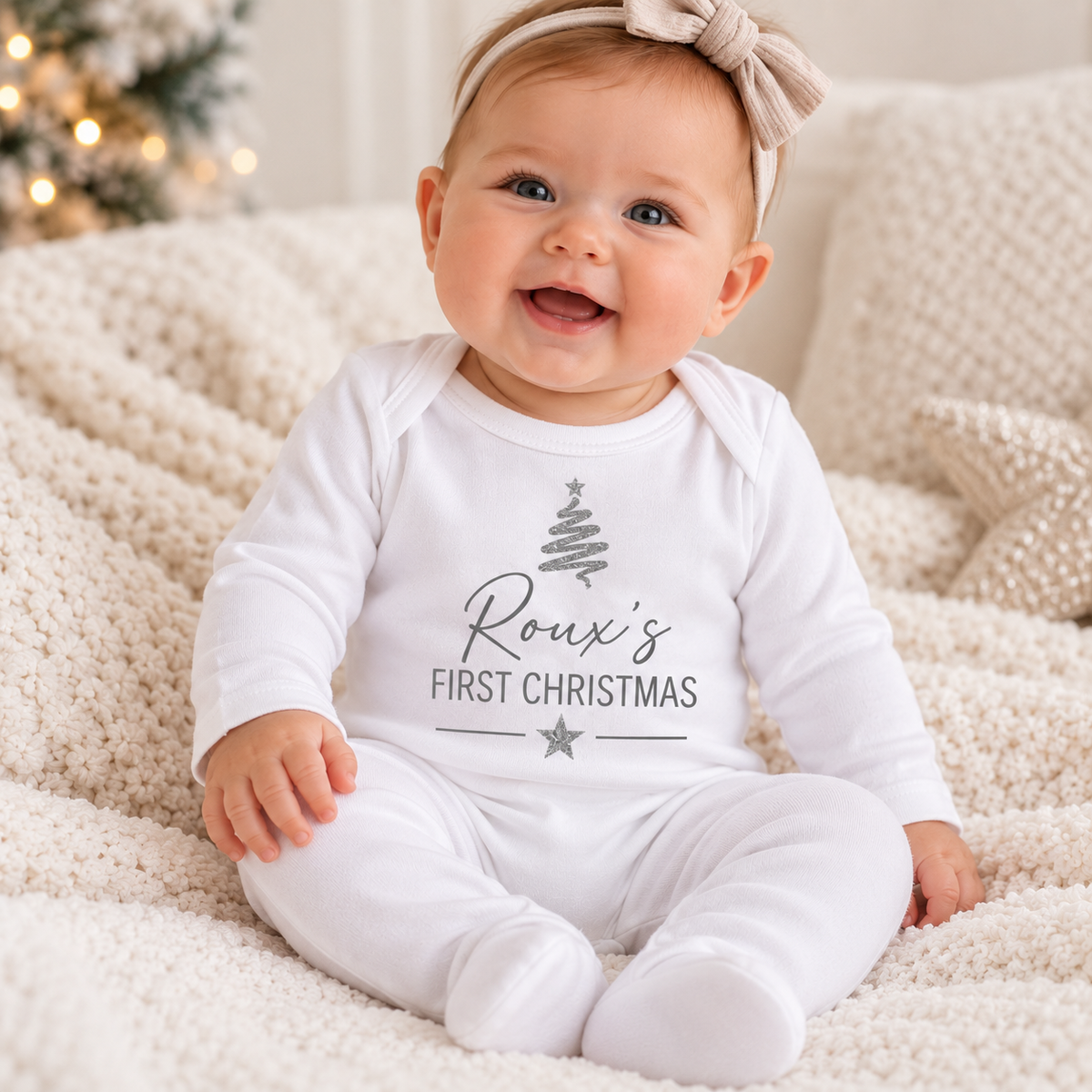 Baby sitting on a cream knitted blanket, wearing a white First Christmas baby grow with a silver tree design, Grace & Grayson.