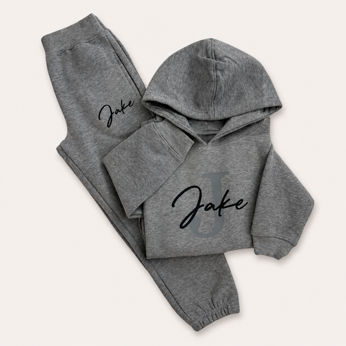 Grey folded hoodie tracksuit with a grey initial and black script name personalisation, laid flat on a cream background. Grace & Grayson