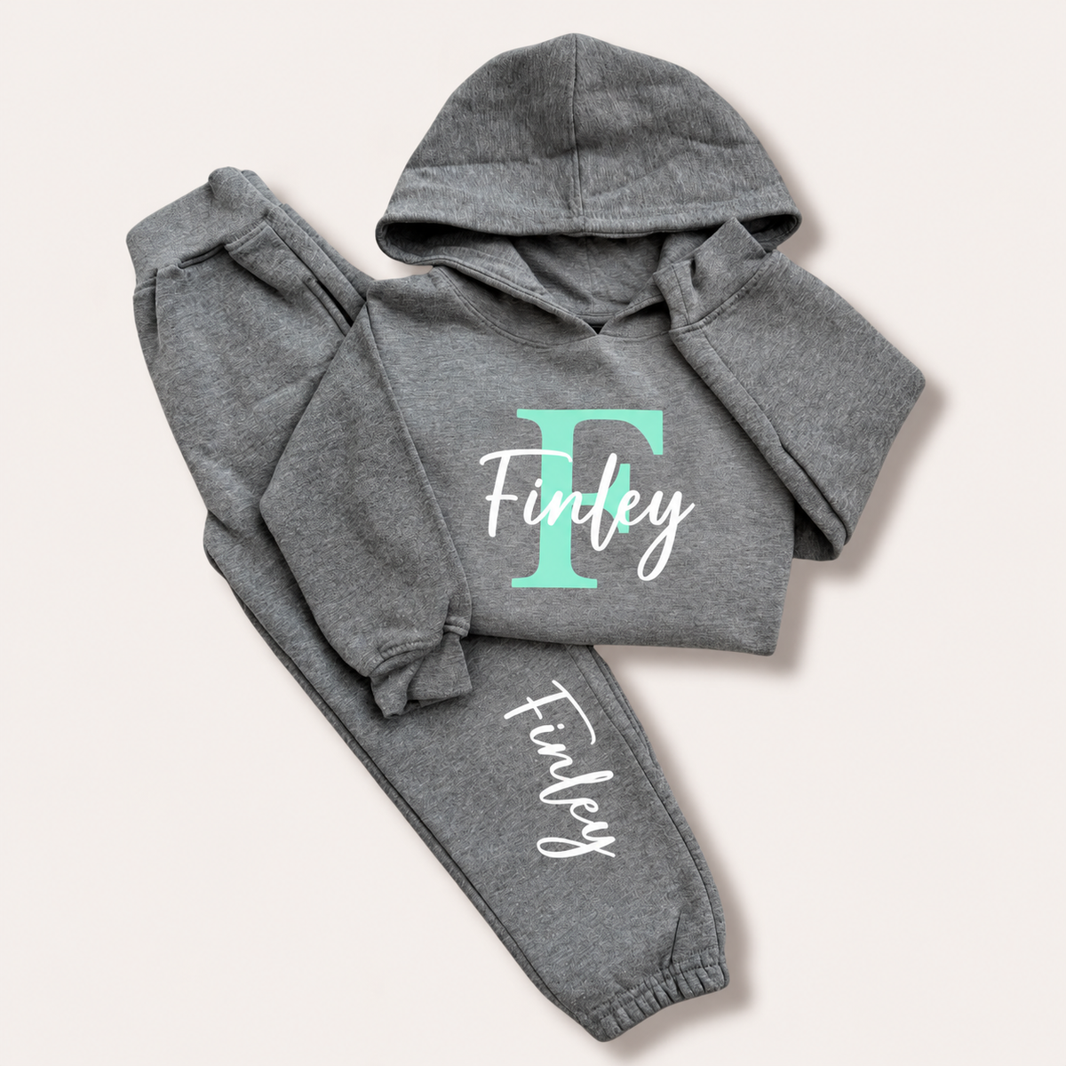 Grey hoodie tracksuit with a mint green initial and white script name personalisation, laid flat on a cream background. Grace & Grayson