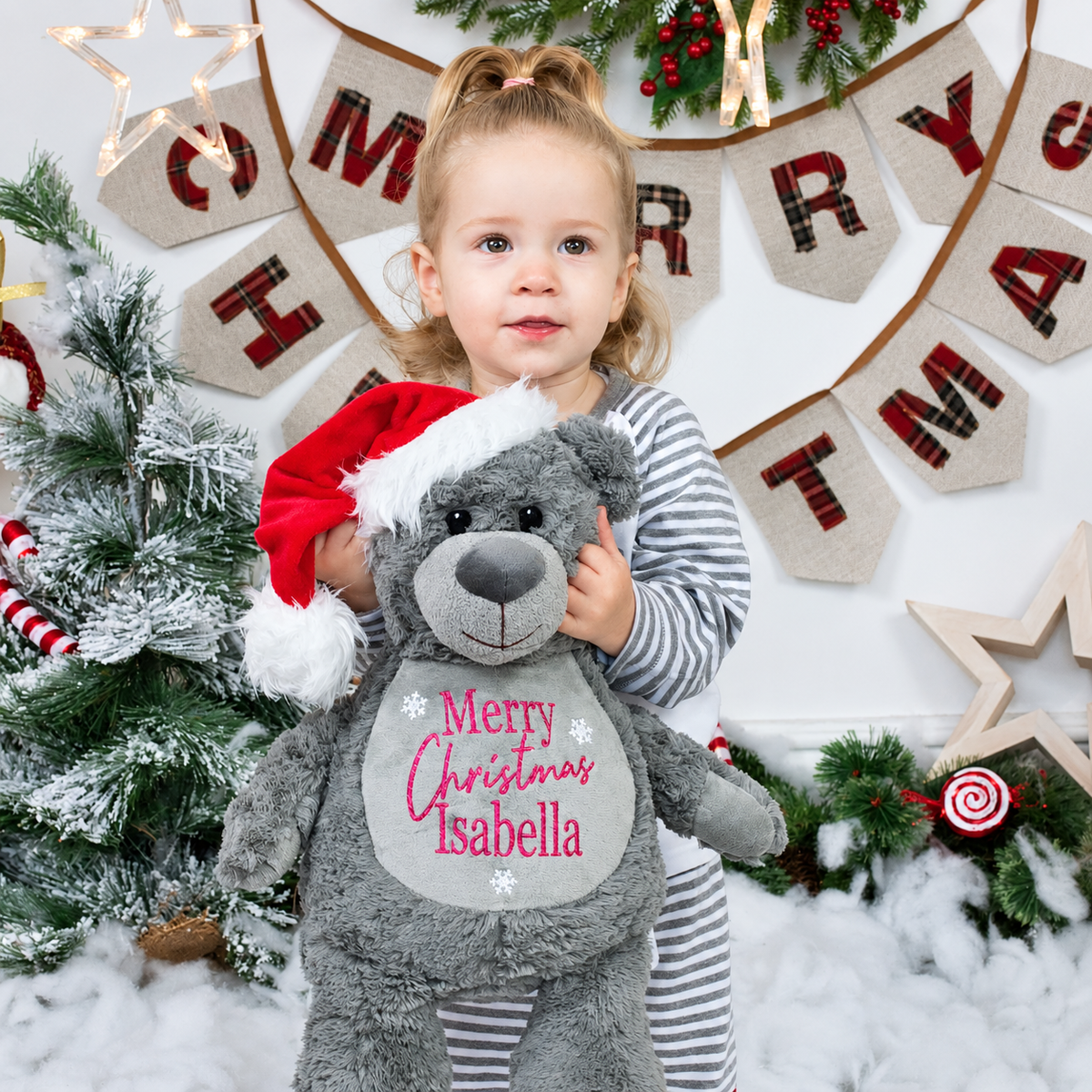 Child holding a grey personalised Christmas teddy bear wearing a Santa hat, in a festive Christmas setting, Grace & Grayson
