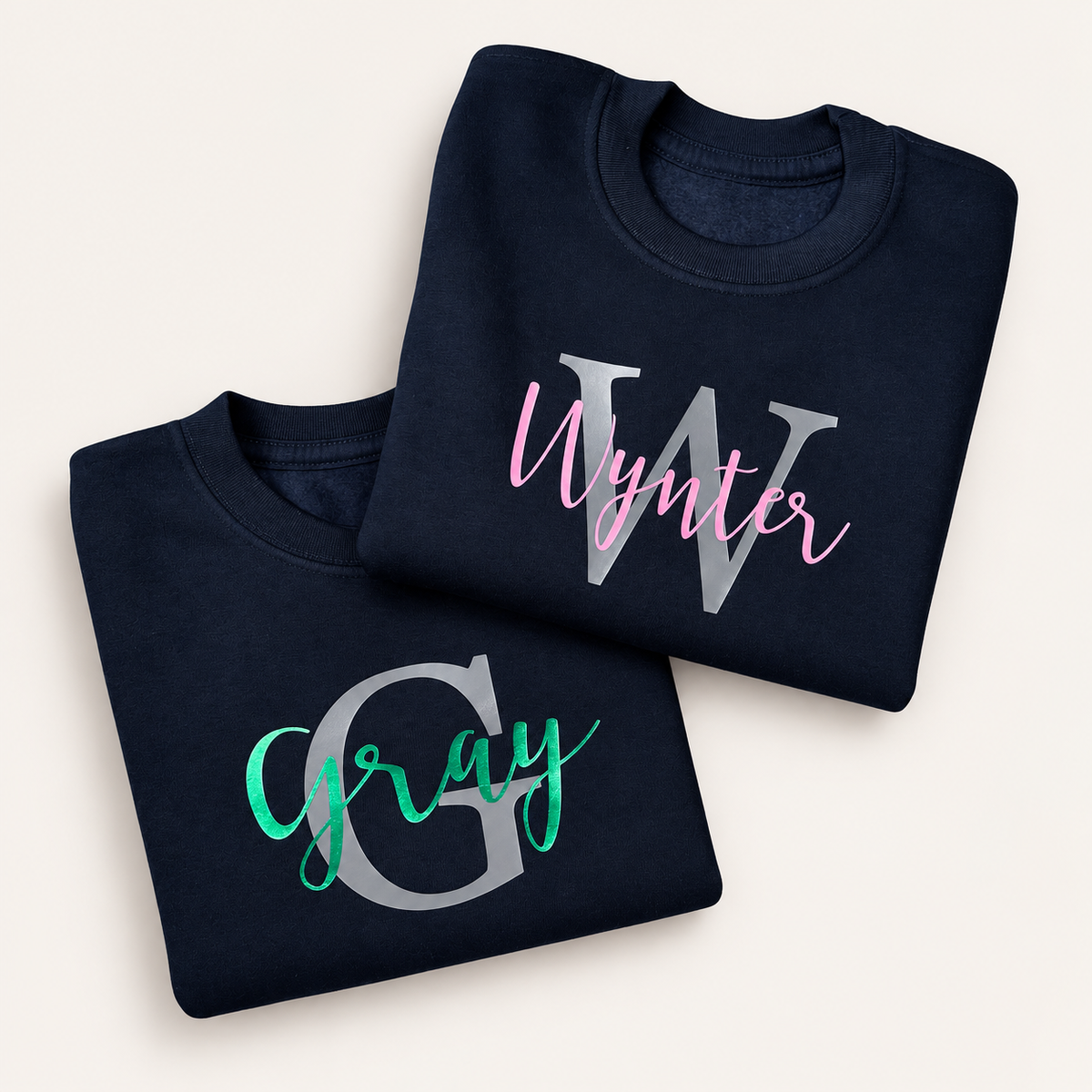 Two folded navy personalised sweatshirts with grey initials and pink and green script names | Grace & Grayson