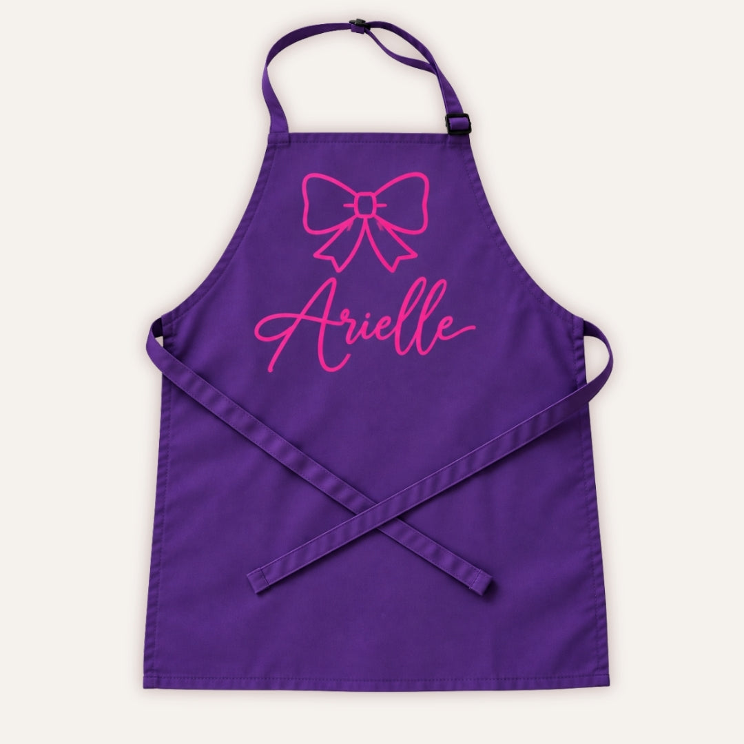 Purple personalised children’s apron with pink bow design on a white background, Grace & Grayson