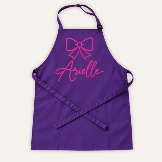Purple personalised children’s apron with pink bow design on a white background, Grace & Grayson