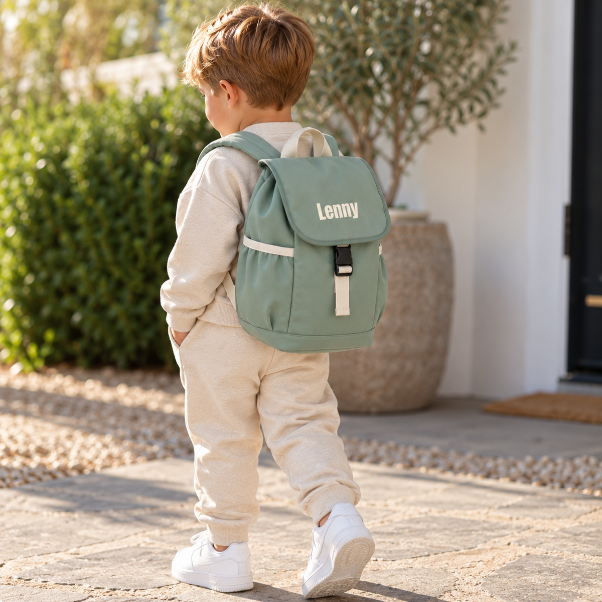 Child walking outdoors wearing a personalised dusty green backpack with cream lettering and straps, Grace & Grayson.