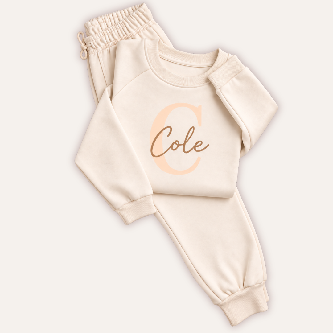 Oat-coloured personalised children’s tracksuit laid flat on a light background, with a large initial and matching name on the sweatshirt and joggers.